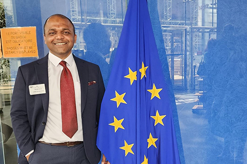 Aayush Varma, a smiling South Asian man in a suit, standing smiling next to a standing EU flag, in front of a blue tinted window.