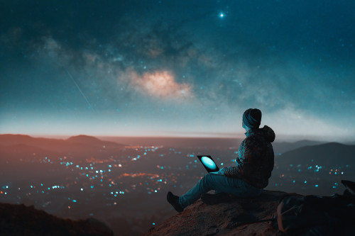 Man with laptop viewing a city from a hill top beneath a star filled sky.