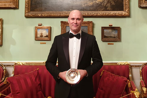 A man dressed in a tuxedo and black bowtie holds a silver plate in front of a lavishly decorated wall.