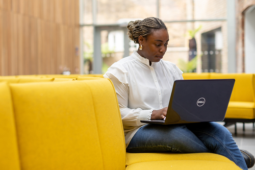 Black female student working with laptop. Sat on a bright yellow sofa on campus.