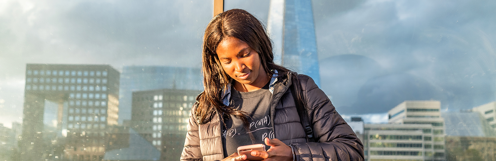 Black female student looking at smartphone, London skyline reflected in the window behind her.