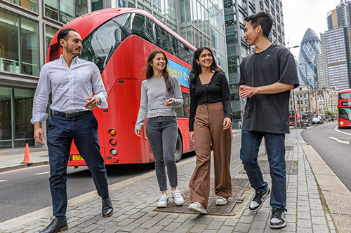 Multicultural student group walking down street in City of London, red London bus and Gherkin building behind.