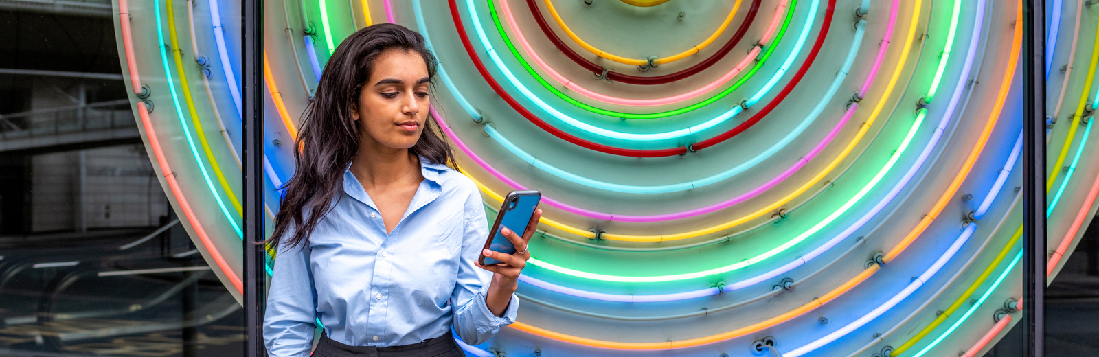 South Asian young woman looking at her phone in front of multicoloured lights near Barbican.