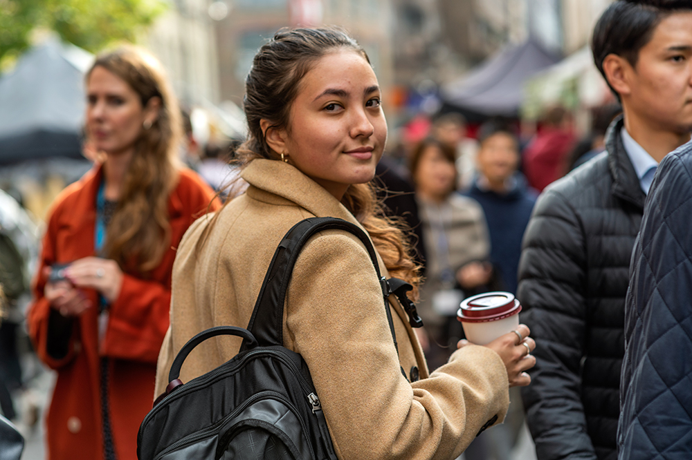 Young female student holds a coffee cup and looks behind at the camera in a busy street